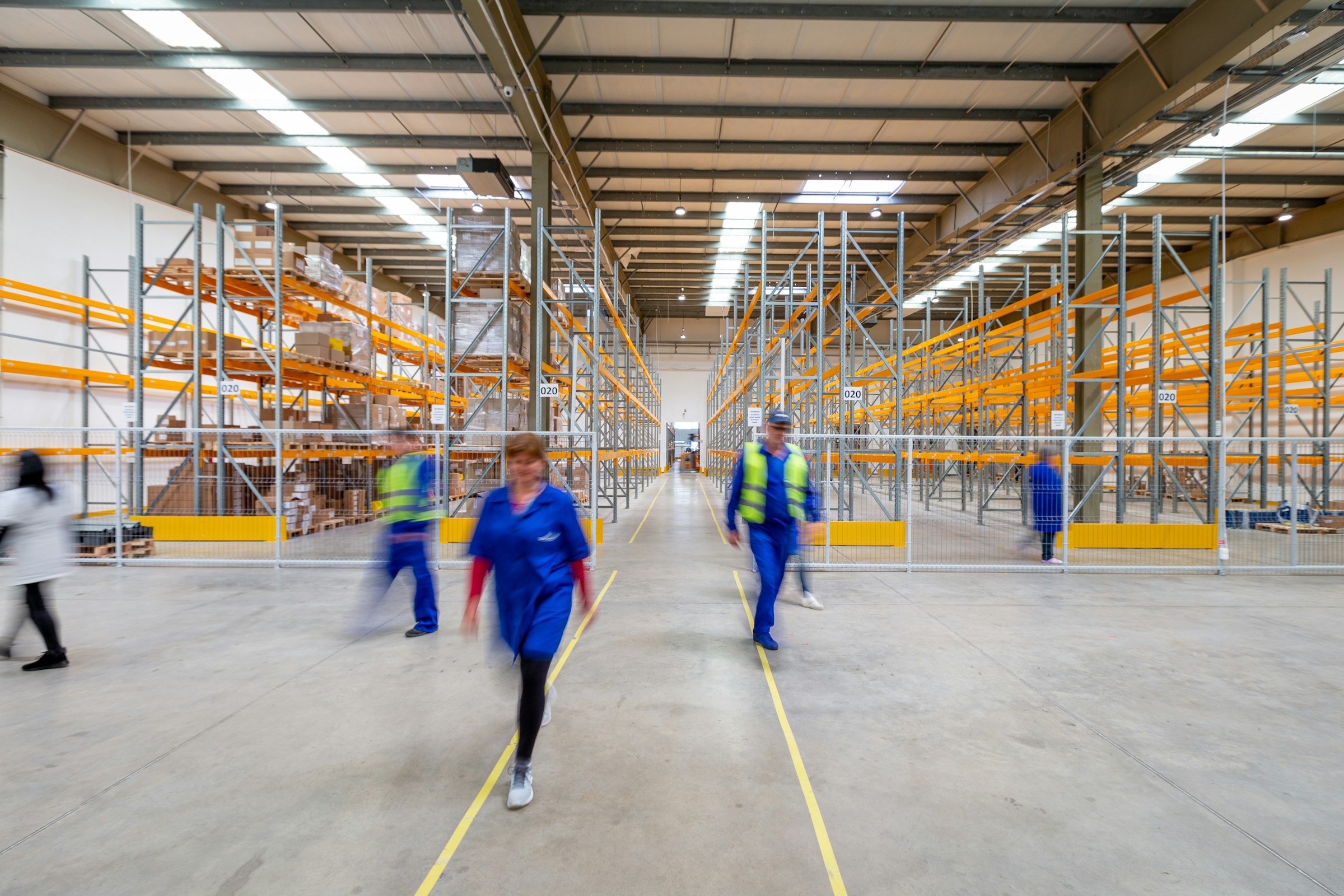 Frontline warehouse workers in hi-vis vests and overalls hurrying down a busy warehouse aisle, illustrating high-volume recruiting challenges