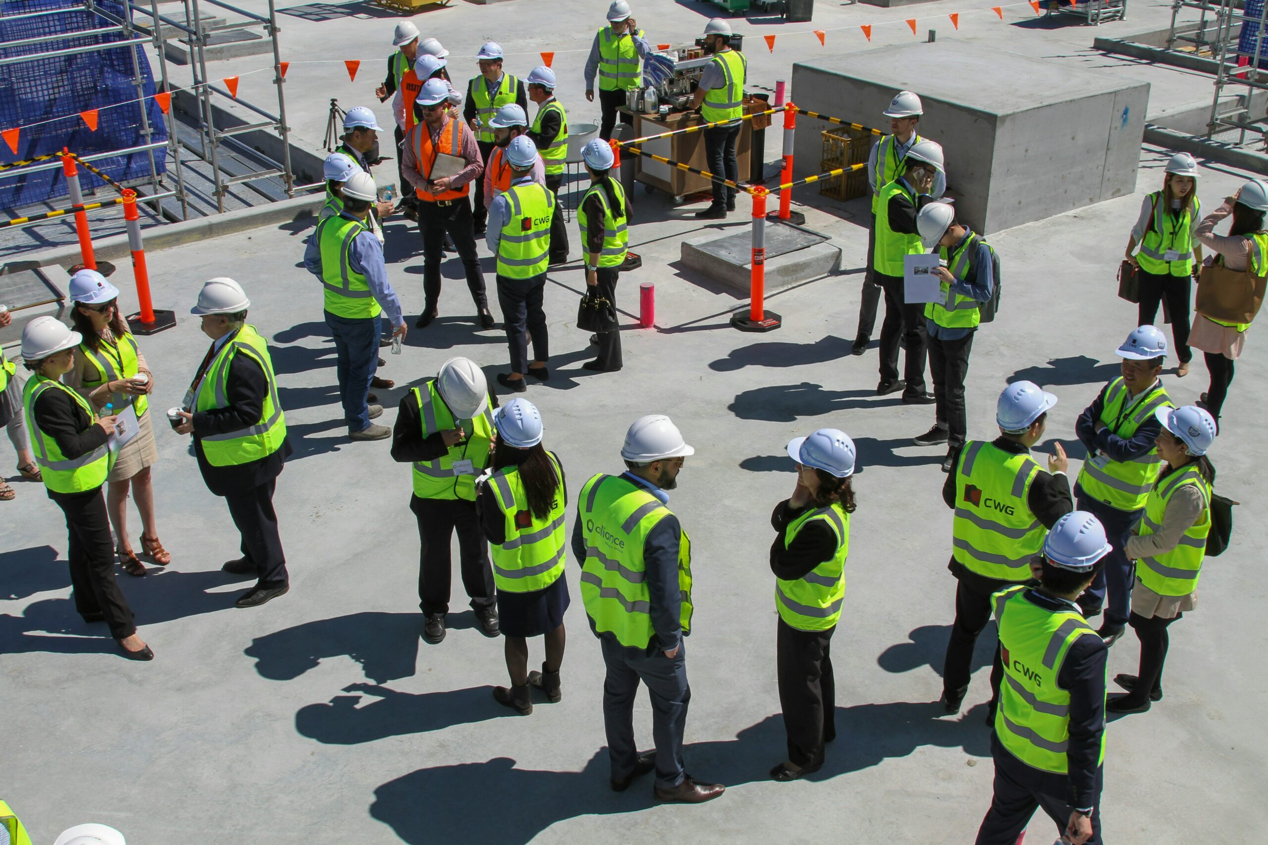 A large team of construction workers in hard hats and hi-vis vests gathers for a briefing on a building site